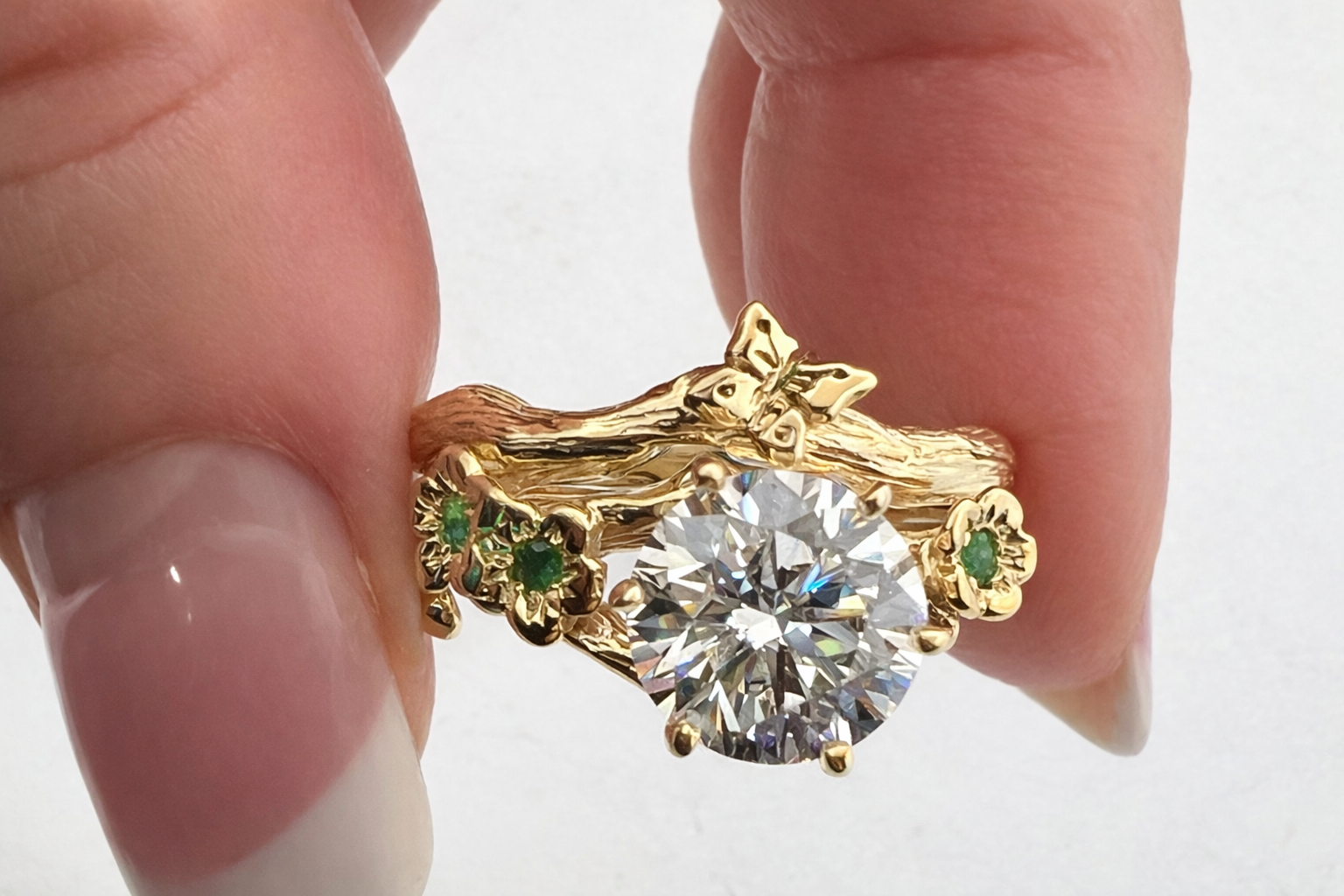 Nature-inspired moissanite engagement ring and gold butterfly band held in fingers, handcrafted bud branch bridal set with emeralds by Teri Lee Jewelry