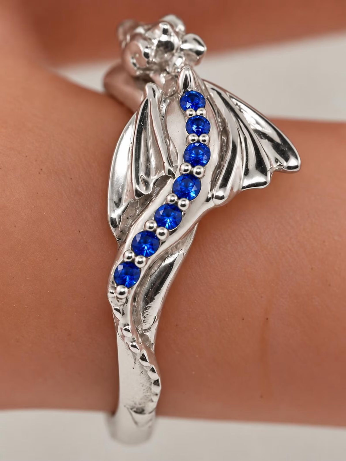 Sterling silver Baby Dragon ring with natural blue sapphires along its back, shown on a finger with sculpted wings and a curled tail.